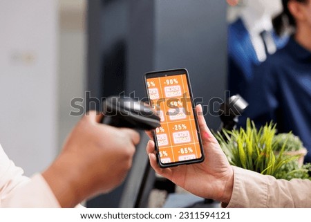 Retail employee scanning smartphone with Black Friday discount coupon while serving customer at cash register desk, close up. Clothing store marketing promotion during seasonal sales Royalty-Free Stock Photo #2311594121