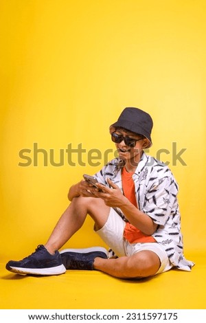 Full length of smiling young Asian  traveler tourist man sit on floor using mobile phone booking hotel taxi isolated on yellow background. Passenger travel on weekend. Air flight journey concept