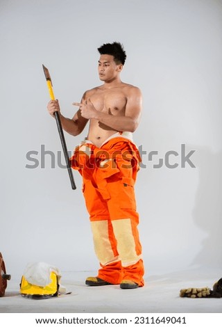 Topless firefighter stands wielding an iron axe in one hand while using the other to point directly at it this confident gesture accentuates his strength.