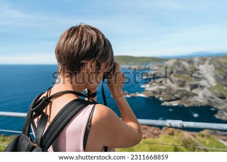 A Caucasian tourist captures Ireland's breathtaking beauty as she photographs lush green cliffs bathed in enchanting light, under a serene azure sky and against the backdrop of the deep blue sea.