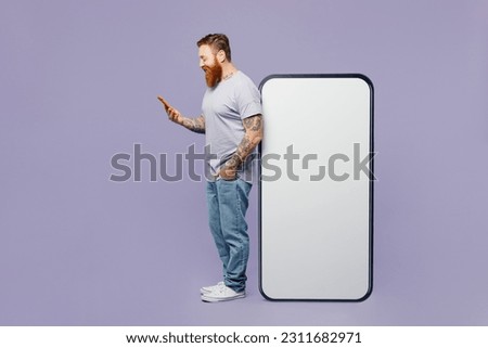 Full body side view fun young redhead man wear violet t-shirt casual clothes big huge blank screen mobile cell phone with copy space mockup area using smartphone isolated on plain purple background
