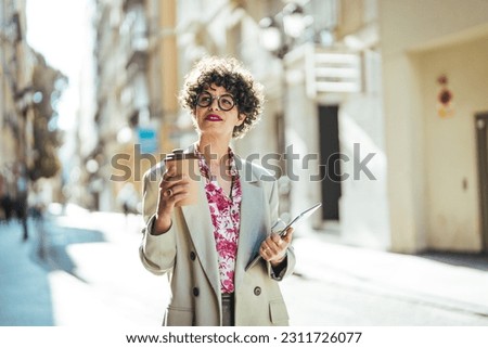 Female model with natural black hair dressed in formal wear holds touchpad notebook and cup of takeaway coffee thinks about content publication for sharing to website poses outdoor during sunny day