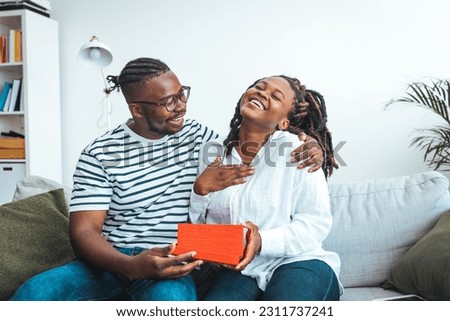 Man presenting gift to his beloved woman at home. Valentine's day celebration. Man surprising his girlfriend with a gift on the couch at home in the living room. 