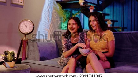 Laughing two Indian Girlfriend Sitting on sofa enjoying late night watching funny TV series at home. Female couple looking comedy movie on television eating popcorn snacks spend weekend time together