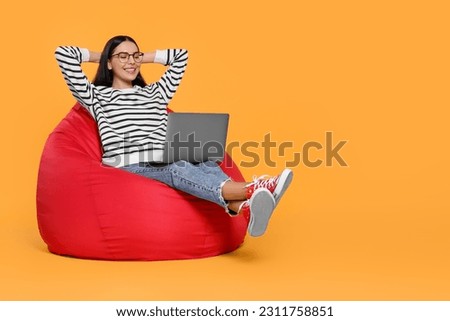 Happy woman with laptop sitting on beanbag chair against orange background