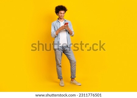 Full size photo of cheerful satisfied guy wear jeans jacket pants shoes look at smartphone write email isolated on yellow color background