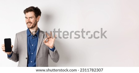 E-commerce and online shopping concept. Smiling businessman show okay sign and empty smartphone screen, demonstrate his account, standing on white background.