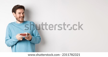 Excited man receive birthday surprise, holding bday cake and smiling happy, standing over white background, making wish on lit candle.