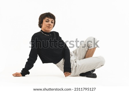 Full shot of a woman in a black jumper and white trousers sitting with her legs crossed and leaning one hand on the ground. High quality photo