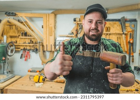 Smiling carpenter holding a hammer makes thumbs up sign in a woodworking studio. High quality photo
