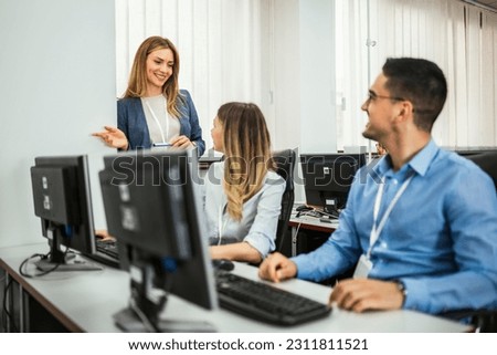 A group of students each sit in front of a desktop in a computer lab. They are each focused on their screens as their female teacher makes her way around to check on each student individually. Royalty-Free Stock Photo #2311811521