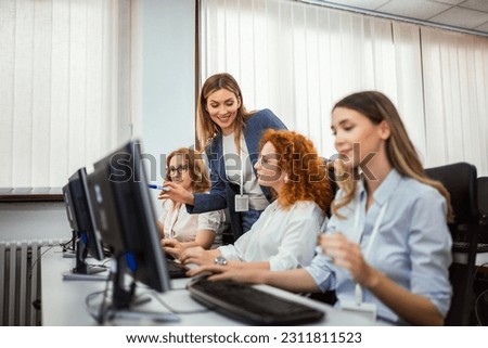 A group of students each sit in front of a desktop in a computer lab. They are each focused on their screens as their female teacher makes her way around to check on each student individually. Royalty-Free Stock Photo #2311811523
