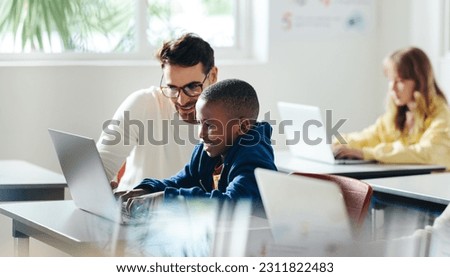 Male teacher helps a young boy with computer-based learning in a classroom setting. Child tutor providing a lesson in an elementary school, with a focus on coding and basic digital literacy. Royalty-Free Stock Photo #2311822483