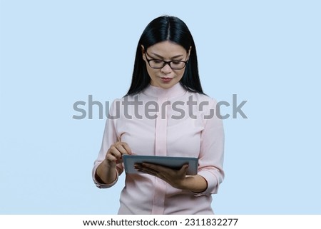 Young smart asian woman in glasses is using a tablet pc. Looking down and scrolling. Isolated over pale blue background.