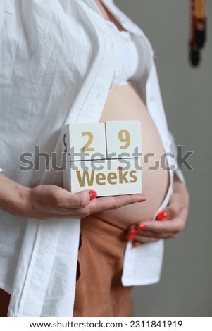 Cropped image of Unrecognizable pregnant woman in unbuttoned white shirt hugging belly and holding cubes with 29 weeks.. Baby care and health. Isolated on a grey background indoor. Space for text.
