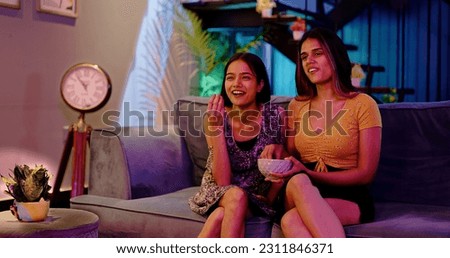 Laughing two Indian Girlfriend Sitting on sofa enjoying late night watching funny TV series at home. Female couple looking comedy movie on television eating popcorn snacks spend weekend time together Royalty-Free Stock Photo #2311846371