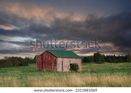 Old wooden, red shed in the middle of a field with a Canadian flag.  Stormy sunset clouds hang heavy as the weathered shed with green roof stands against the storm. 