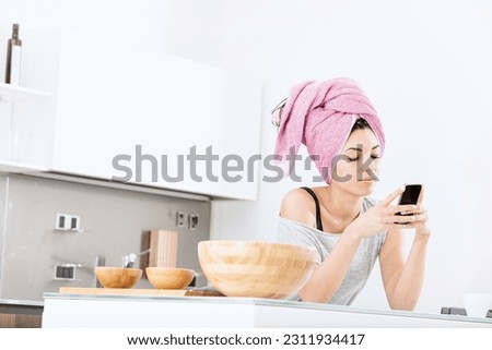 A young woman with a towel on her head, drying her hair, relaxes in her bright and spacious home, scrolling through gossip and social networks on her smartphone. She is happy and content