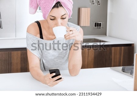 With a towel on her head, a young woman dries her hair while enjoying leisure time in her bright and expansive home. She finds happiness and contentment as she engages with gossip and social networks 