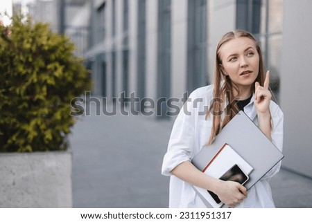 Blonde businesswoman with laptop and book in hand, have idea. point finger up, wow emotions outside office background. Concept of idea and brainstorm. Woman have new ideas or search solutions.