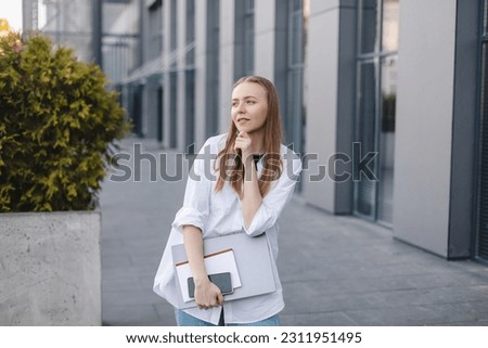 Blonde businesswoman with laptop and book in hand, happy dreaming look and finger to chin outside office background. Concept of idea and brainstorm. Woman think about new ideas or search solutions.