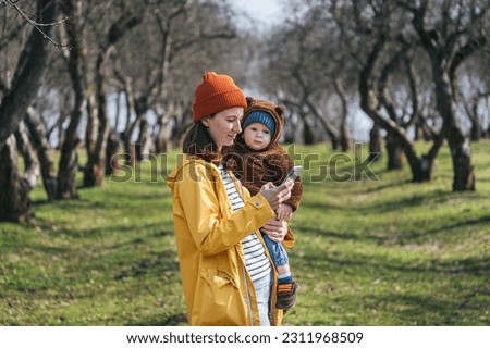 a young mother stands in nature with a child and looks at the phone screen against the background of an alley with trees without leaves
