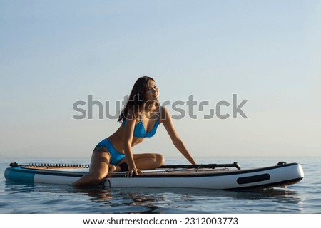Asian beautiful girl in a blue bikini is engaged in water sports. She sits on a surfboard and laughs. in the ocean against the backdrop of sunset. Woman on paddleboarding , Sup.