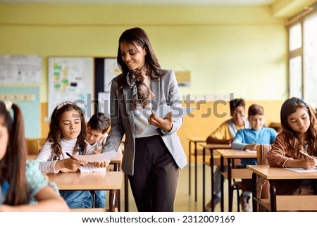 Happy Hispanic teacher giving exam paper to her student during a class at primary school. Royalty-Free Stock Photo #2312009169
