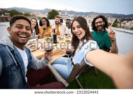 Group of diverse friends having fun at rooftop party. Beautiful woman taking selfie at barbecue dinner time. Smiling people eating and drinking on outdoor terrace. Positive friendship relations.