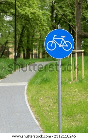 White bicycle lane road traffic sign on blue round background. Bike sign on metal pole, blur narrow street in city park.
