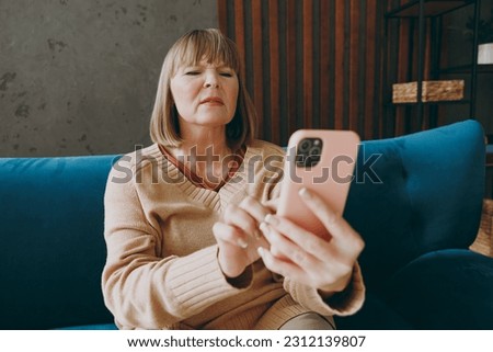Elderly woman 50s years old wears casual clothes sits on blue sofa hold use mobile cell phone without glasses squint stay at home flat rest relax spend free spare time in living room indoor grey wall Royalty-Free Stock Photo #2312139807