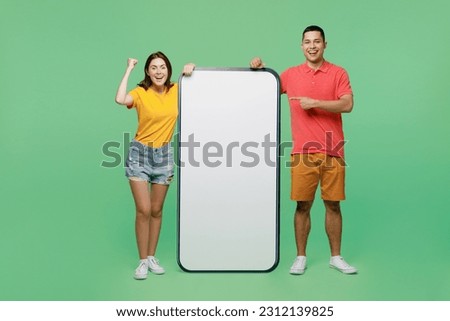 Full size fun young couple two friends family man woman wear t-shirts together near big huge blank screen mobile cell phone with area do winner gesture isolated on pastel plain light green background