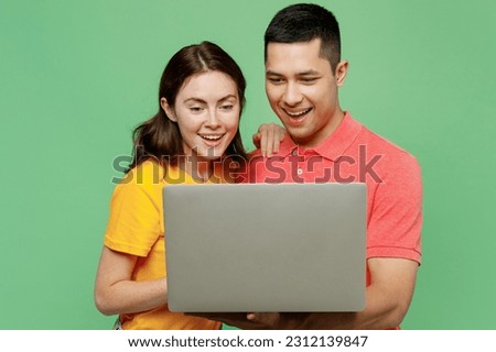 Young smiling happy cheerful fun IT couple two friends family man woman wear basic t-shirts together hold use work on laptop pc computer isolated on pastel plain light green background studio portrait