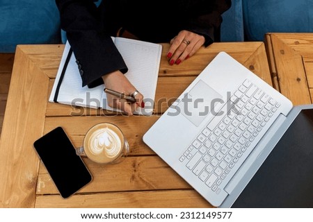Female hands with red manicure on the background of a white laptop. The girl in the cafe works on a laptop. On the table is a cup of coffee, a diary and a phone next to a laptop