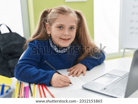 Adorable blonde girl student using laptop writing notes at classroom