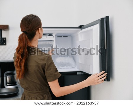A woman in the kitchen of her home opened an empty freezer with ice inside, home refrigerator, defrosted, view from the back. Royalty-Free Stock Photo #2312163937