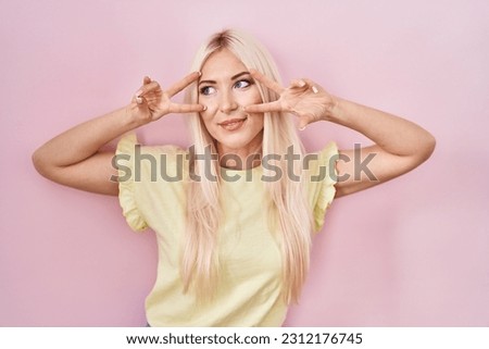 Caucasian woman standing over pink background doing peace symbol with fingers over face, smiling cheerful showing victory 