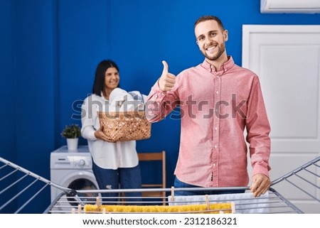 Young couple hanging clothes at clothesline smiling happy and positive, thumb up doing excellent and approval sign 