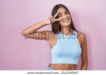 Young brunette woman standing over pink background doing peace symbol with fingers over face, smiling cheerful showing victory 
