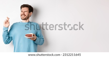 Celebration and holidays concept. Young man celebrating birthday, cross fingers for good luck, making wish on bday cake with lit candle, standing over white background.