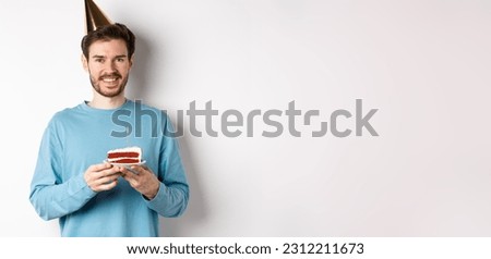 Celebration and holidays concept. Happy young man in party hat celebrating birthday, holding bday cake and smiling, standing over white background.