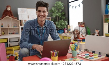 Young hispanic man preschool teacher using laptop sitting on table at kindergarten Royalty-Free Stock Photo #2312234573