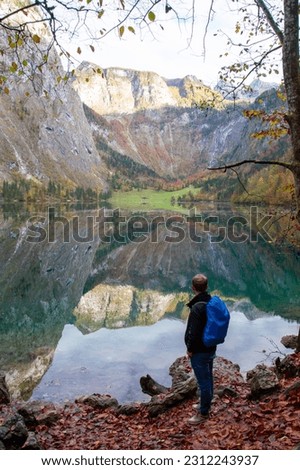 The picture shows a man standing in front of a tranquil alpine lake on a beautiful autumn day. The mighty mountains of the Bavarian Alps reflect beautifully in the water of the lake.
