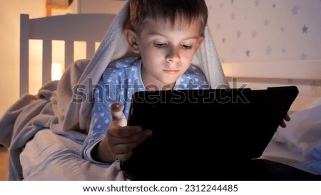 Portrait of cute boy in pajamas lying in bed and browsing internet on tablet computer. Children education, development, kids using gadgets secrecy, privacy.
