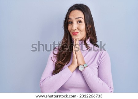 Young brunette woman standing over blue background praying with hands together asking for forgiveness smiling confident. 