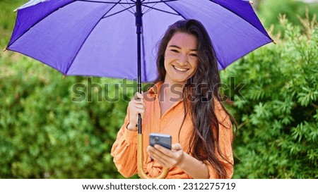 Young beautiful hispanic woman holding umbrella using smartphone at park