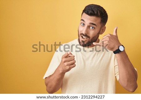 Handsome hispanic man standing over yellow background smiling doing talking on the telephone gesture and pointing to you. call me.  Royalty-Free Stock Photo #2312294227
