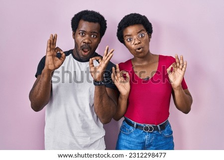 Young african american couple standing over pink background looking surprised and shocked doing ok approval symbol with fingers. crazy expression 