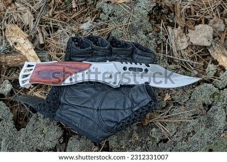 one new iron beautiful hunting modern folding with a brown handle a sharp knife lies on a black leather mitten with cut off fingers on an old wooden stump on the street in the daytime
