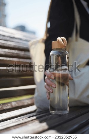 Young woman sitting on a bench in the morning and holding a glass water bottle in hand. Unrecognizable female person chilling outdoor in a sunny day with an eco bottle of fresh water Royalty-Free Stock Photo #2312352955